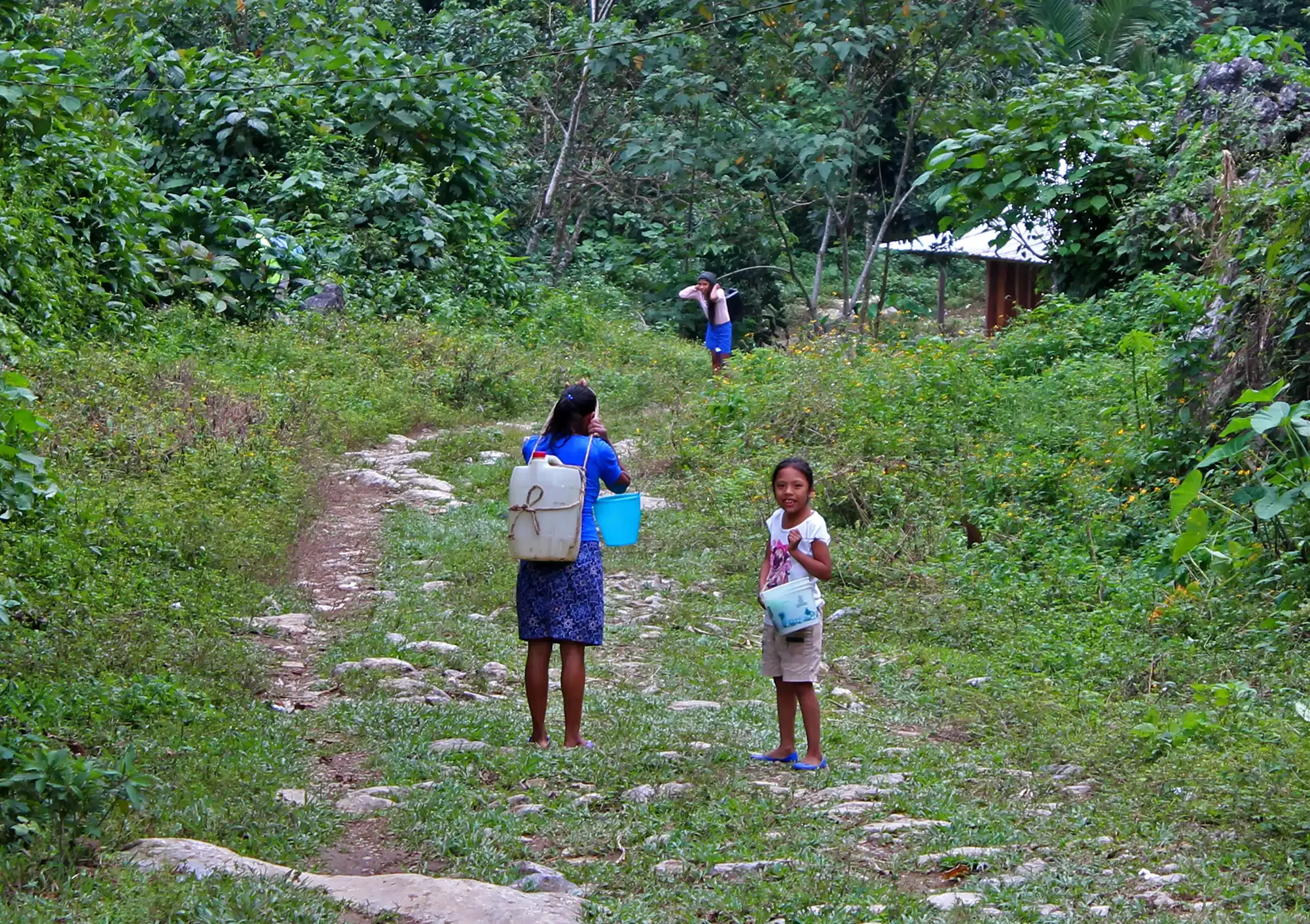 Donate Agua-Vida-2025-Walking for water Donate Agua Vida. A woman and her two little daughters carrying buckets or containers full of water through a path across the mountains in the sierra Mazateca, Oaxaca, Mexico.